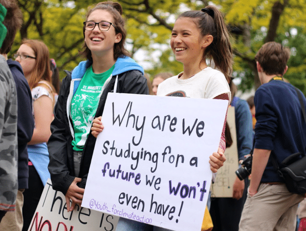 Two young people stand in a crowd holding hand-drawn signs. One sign reads, “Why are we studying for a future we won’t even have!”