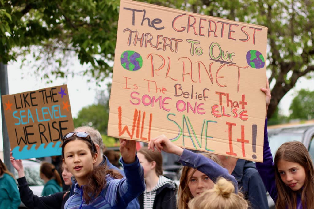Young faces in a crowd hold protest signs. They read, “The greatest threat to our planet is the belief that someone else will save it,” and “Like the sea level, we rise.”